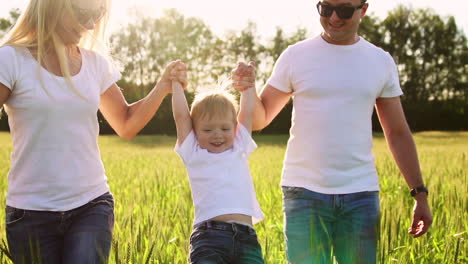 una familia joven con un niño pequeño corriendo en un campo con espigas de trigo balanceando a un niño en sus brazos que ríe y sonríe de felicidad