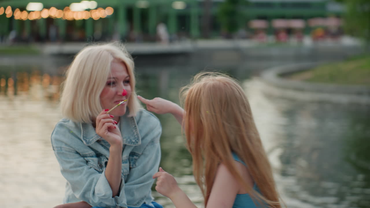 portrait of smiling mom and dancing daughter by lake at sunset with painted faces and brushes, joyful interaction and tender bond, colorful dots on cheeks and noses in warm evening light