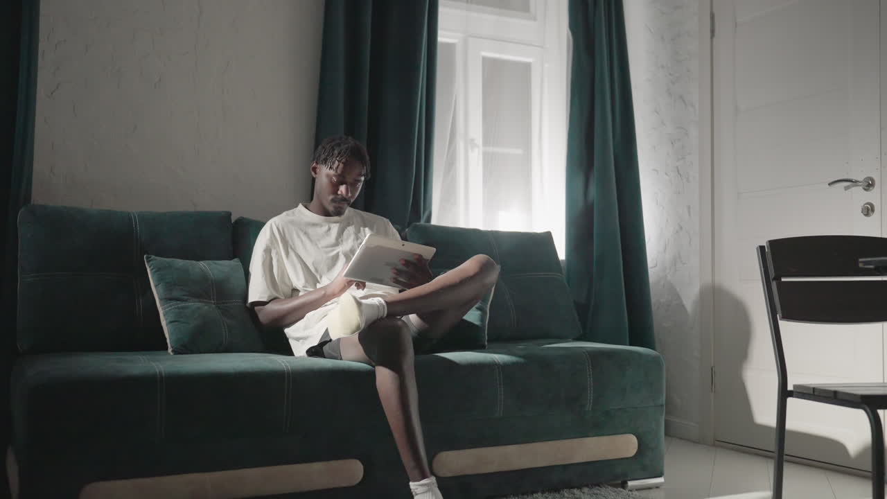 Champ sitting on teal sofa, reading tablet with full attention in cozy, modern living room. Soft light flows through window, creating a peaceful atmosphere with a minimalist design