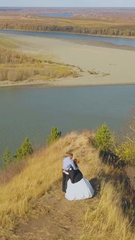 just married couple enjoys view of calm river from steep hill top with dry grass on autumn day aerial view