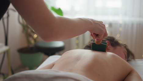 fair skin woman lying on massage table receiving back massage with textured roller from waist to neck in softly lit spa room with blurred white curtain and green plant in background