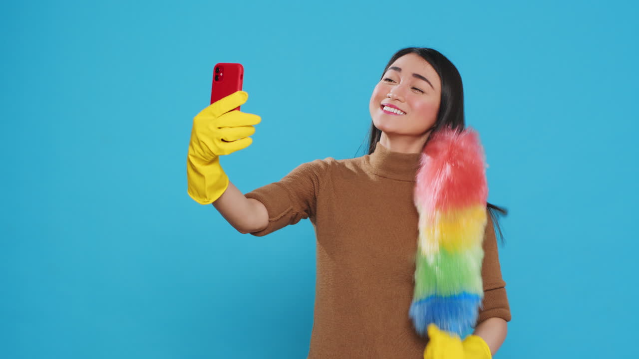 Smiling housekeeper holding colorful feather dust while taking selfie with mobile phone