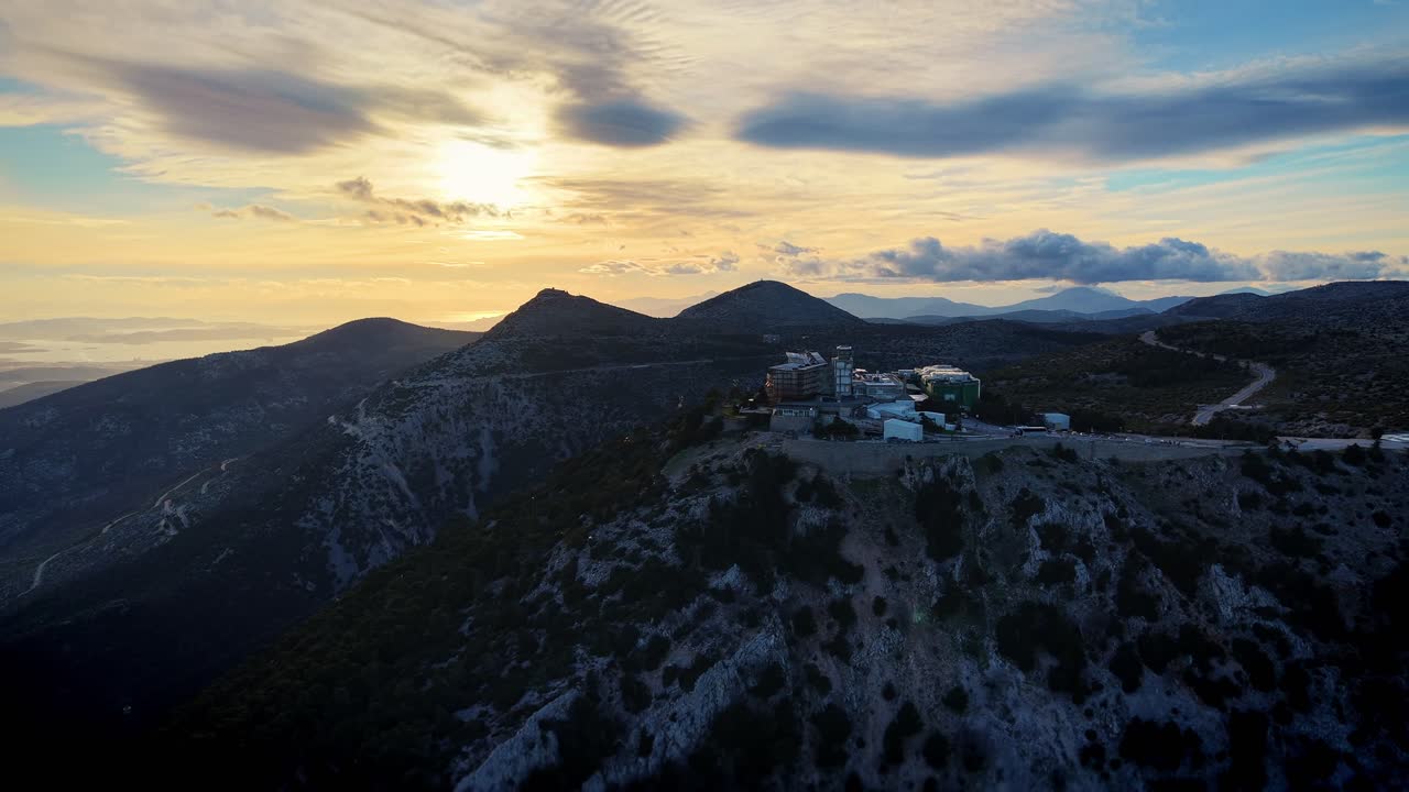 1100 meters altitude, the drone moves rightward capturing Regency Casino Mont Parnes atop its mountain peak in the frame's distance. Jagged mountaintops rise ahead bathed in golden sun