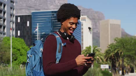 Hombre-Afroamericano-Sonriente-Usando-Un-Teléfono-Inteligente,-Usando-Audífonos-Y-Mochila-En-La-Calle-De-La-Ciudad