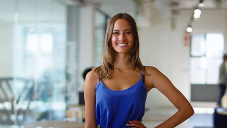 Portrait-Of-Young-Smiling-Businesswoman-Standing-In-Busy-Modern-Open-Plan-Office