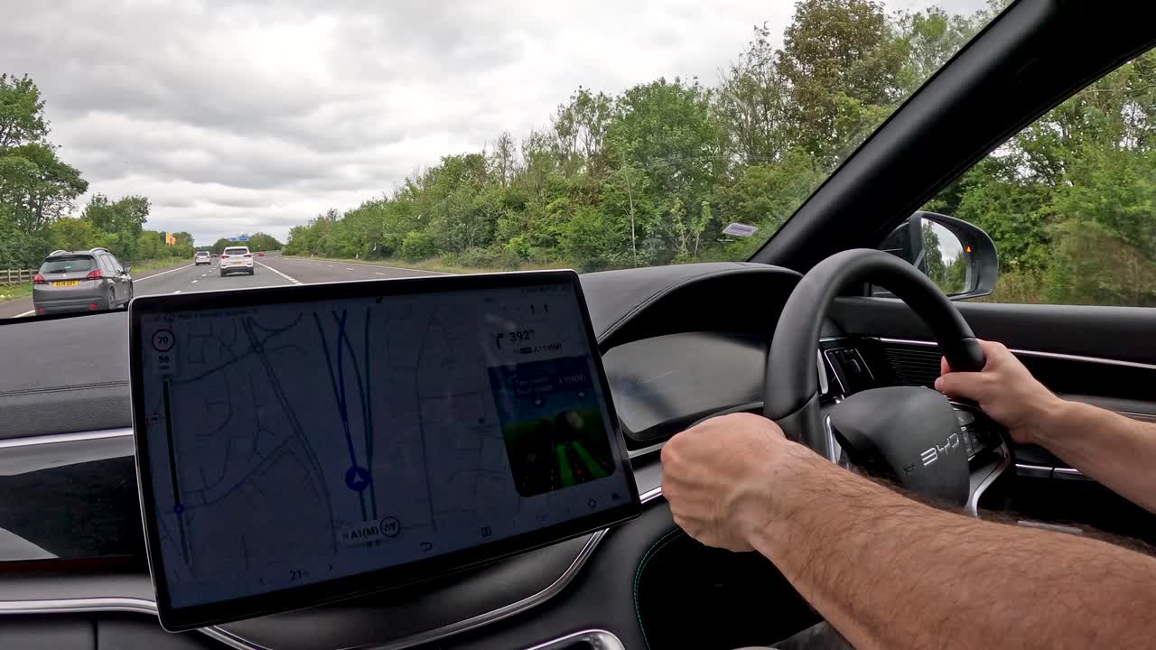 A person operates a modern electric vehicle on a UK motorway, interacting with a large dashboard touchscreen for navigation. Overcast daylight, interior perspective, smooth camera movement