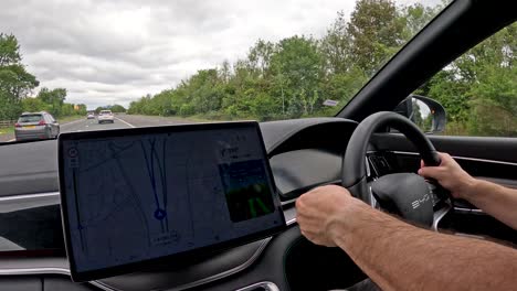 a person operates a modern electric vehicle on a uk motorway, interacting with a large dashboard touchscreen for navigation. overcast daylight, interior perspective, smooth camera movement