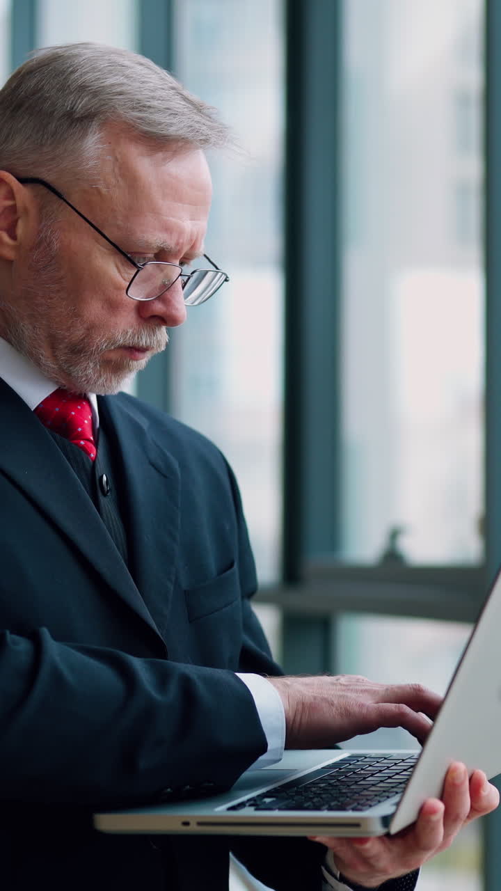 Serious businessman with laptop in hands thinking about future meeting checking presentation standing near window with panoramic city view. Video from the side. Vertical video