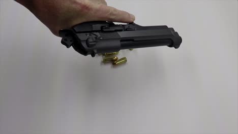 A-person-picking-up-and-cocking-loading-a-black-firearm-with-live-ammunition-rounds-on-a-white-table