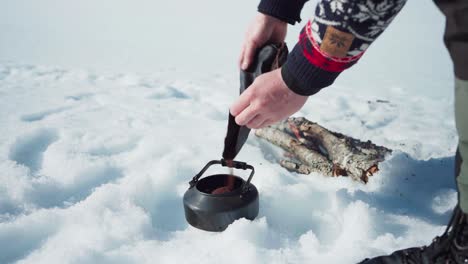 Hombre-Vertiendo-Café-Molido-En-La-Tetera-Cerca-De-La-Leña-En-Llamas-En-La-Nieve