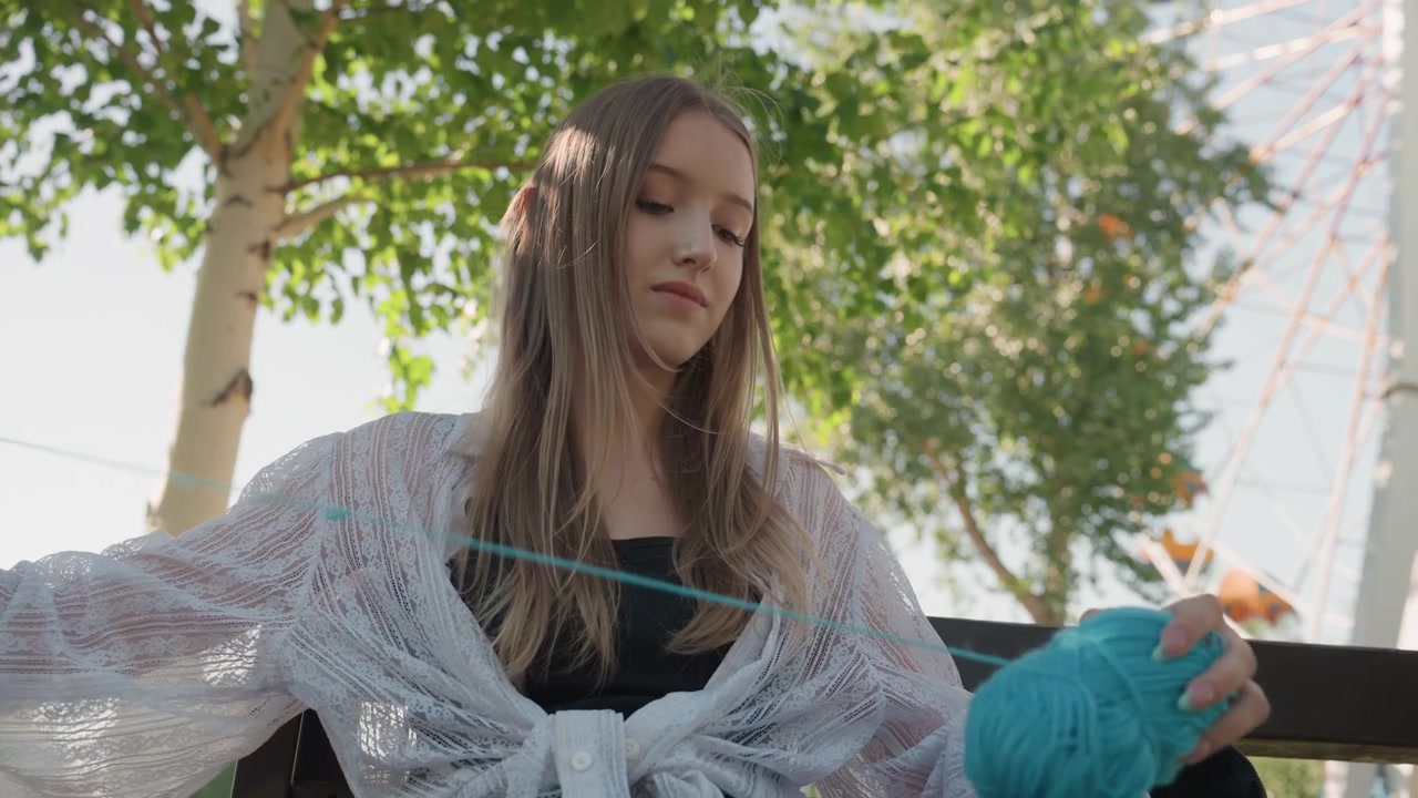 Primo piano di giovane donna che tiene del filato blu e si prepara a lavorare a maglia all'aperto, con alberi e una ruota panoramica sullo sfondo. La donna indossa un top di pizzo bianco sopra una maglietta nera, concentrandosi sulla manipolazione del filato turchese.