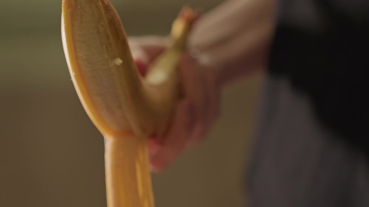 Close up of hands peeling ripe banana from stem to middle. Banana with brown spots is held. Indoor setting with neutral background remains out of focus throughout