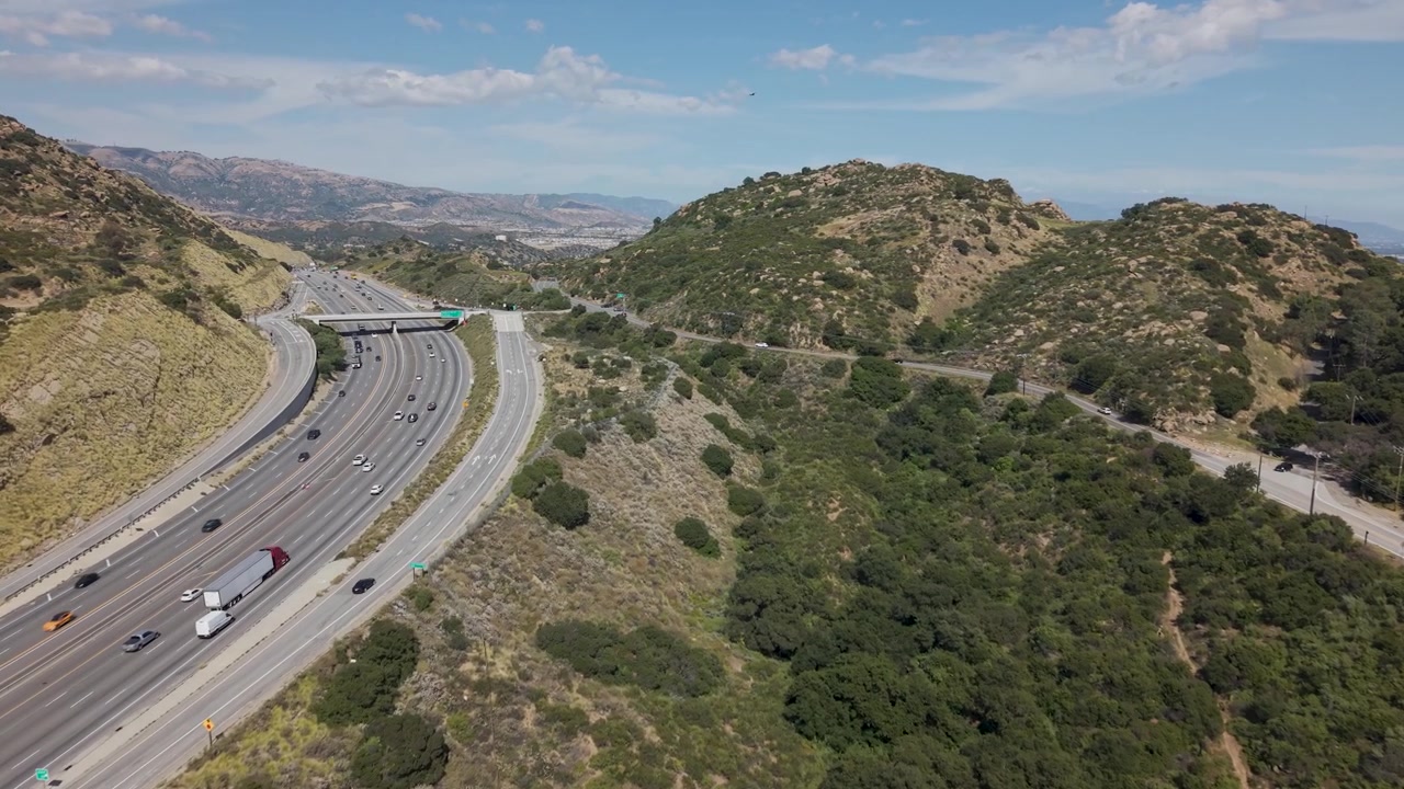 Veduta aerea del traffico che si muove lungo l'autostrada attraverso il Passo di Santa Susana e la Valle di San Fernando, California, con strade tortuose, colline aride, macchie verdi e cielo sereno. Inquadratura ampia