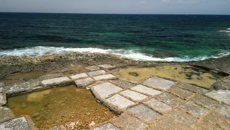 Aerial-footage-revealing-the-sea-and-horizon-with-geometric-salt-flats-and-sea-waves-in-the-foreground-in-Gozo,-Malta