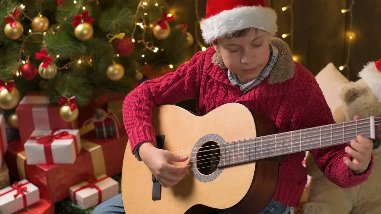 niño adolescente tocando la guitarra, sentado en el interior cerca del árbol de navidad decorado con luces, vestido como ayudante de papá noel - ¡feliz navidad y felices fiestas!