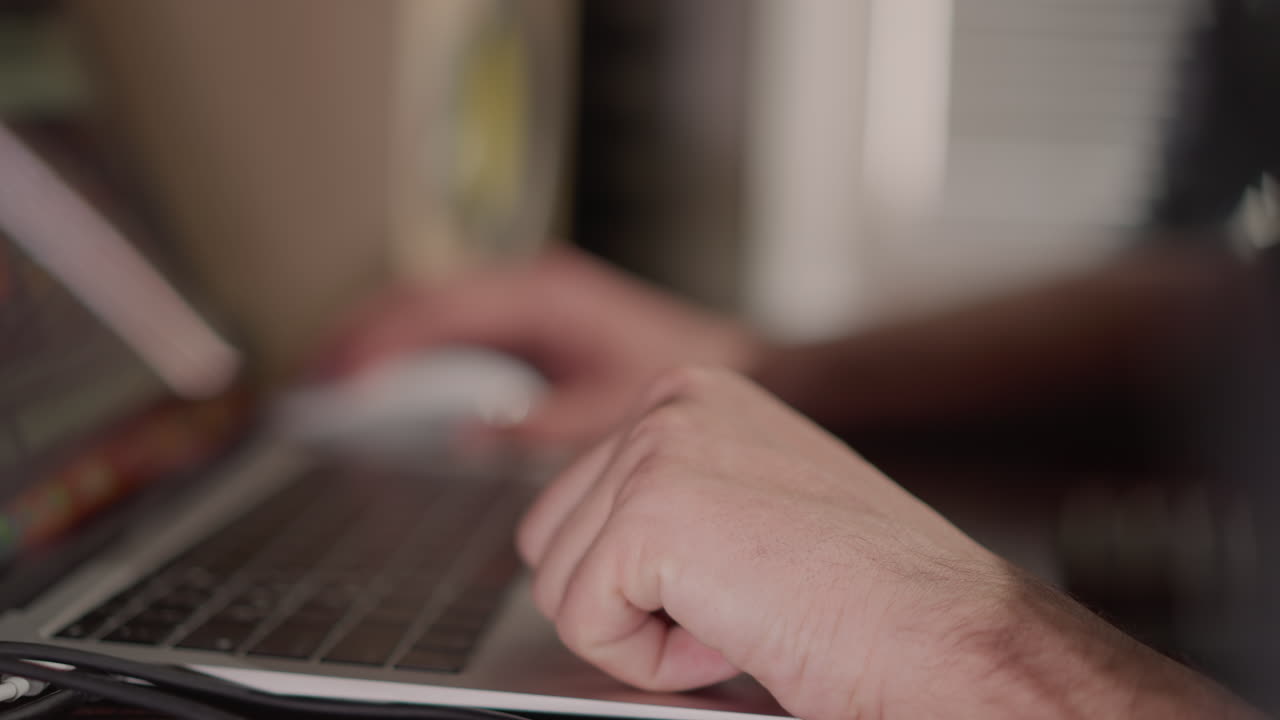 Close-up of hands using a laptop, with one hand typing on the keyboard and the other hand on a mouse. The background is blurred, emphasizing the focus on the interaction with the computer