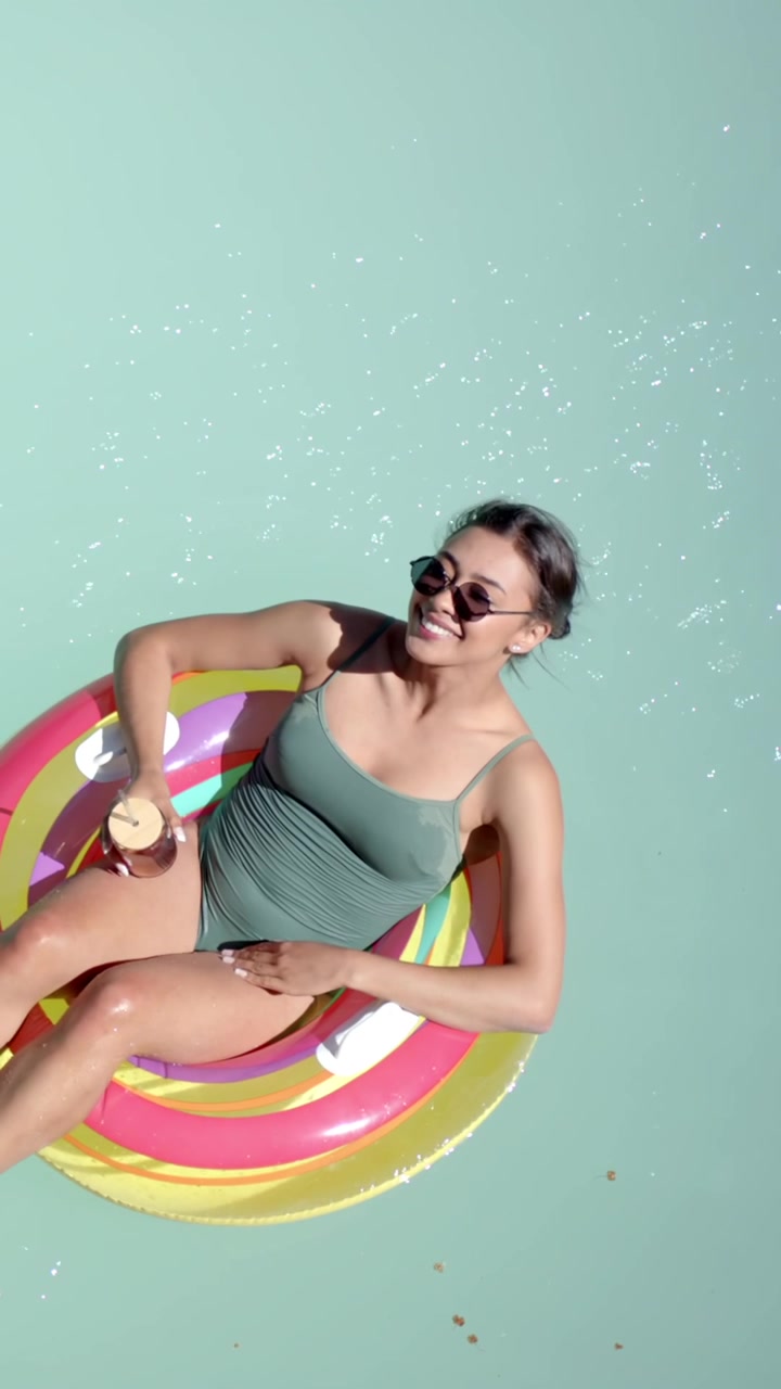 Vertical video: Woman in swimsuit sipping cup on float in pool noticing offframe cue, copy space