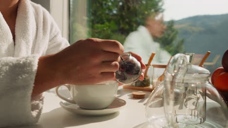 man uses teaspoon taking tea out of glass jar. man tries preparing natural tea in morning at breakfast time. enjoying herbal infusion for good health