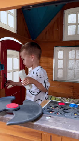 cute caucasian baby boy plays inside the wooden playhouse. kid stands at the toy stove and talks. toddler lives the house and approaches camera. vertical video.