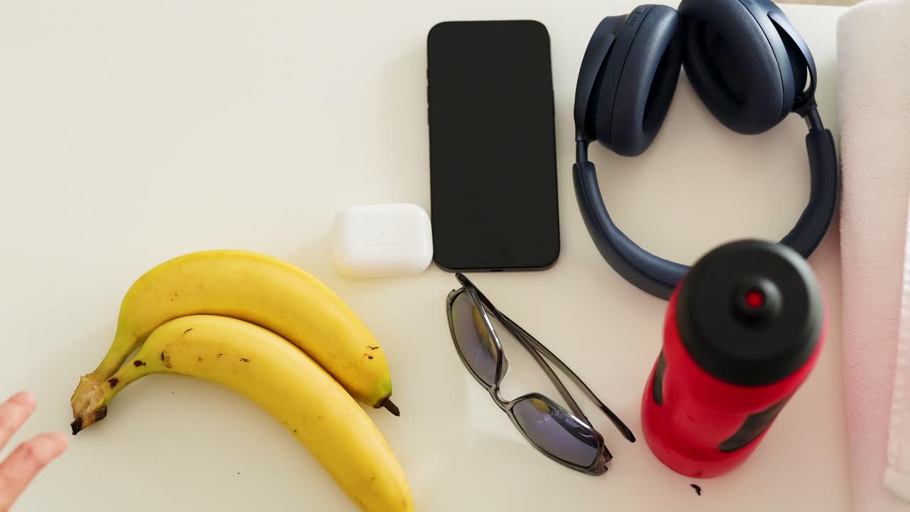 Hands swiftly collect items like a smartphone, headphones, and sunglasses from a table in bright lighting