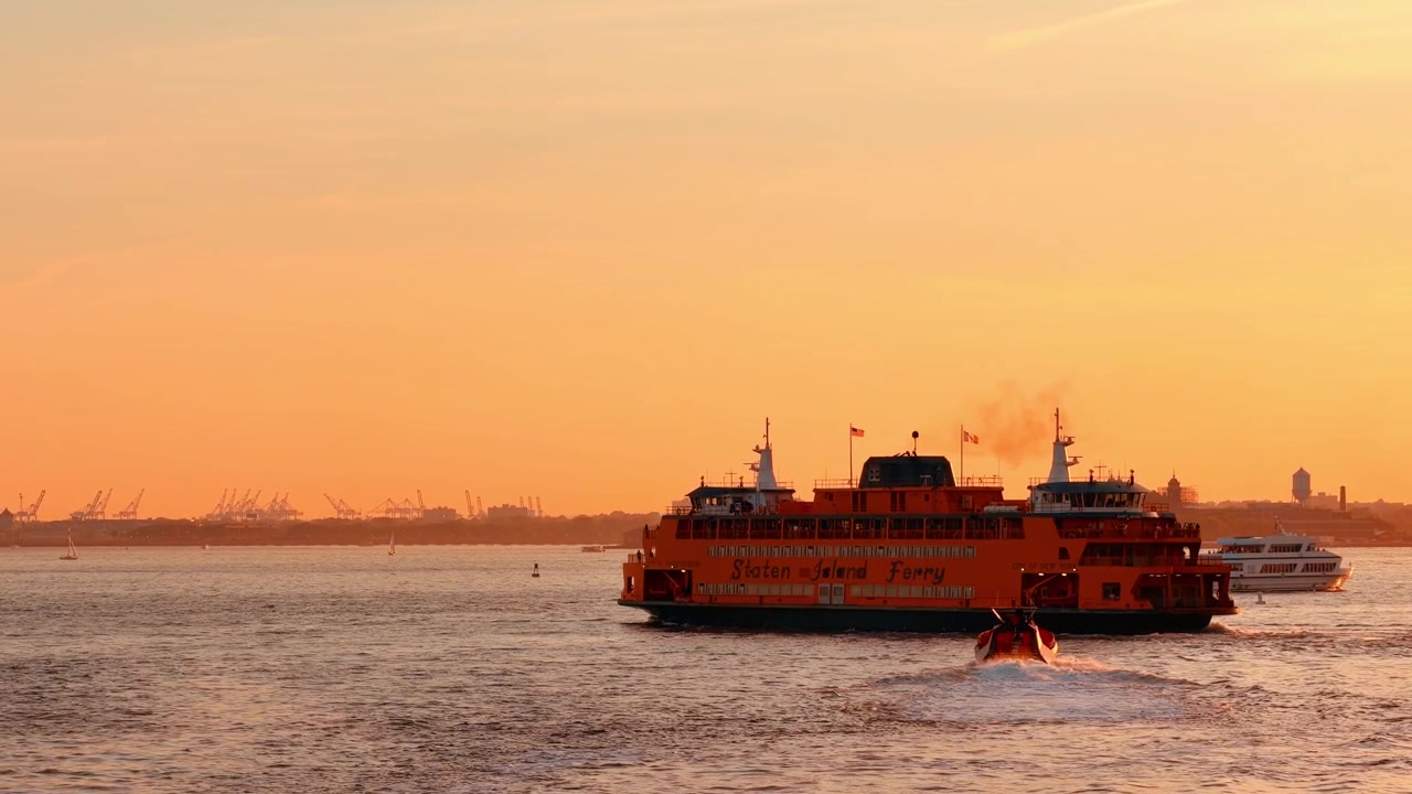 New York, USA, 26. mai 2026: Staten Island-fergen krysser New York Harbor ved solnedgang. Oransje fartøy seiler forbi havnekraner under varm gyllen himmel med disig horisont.