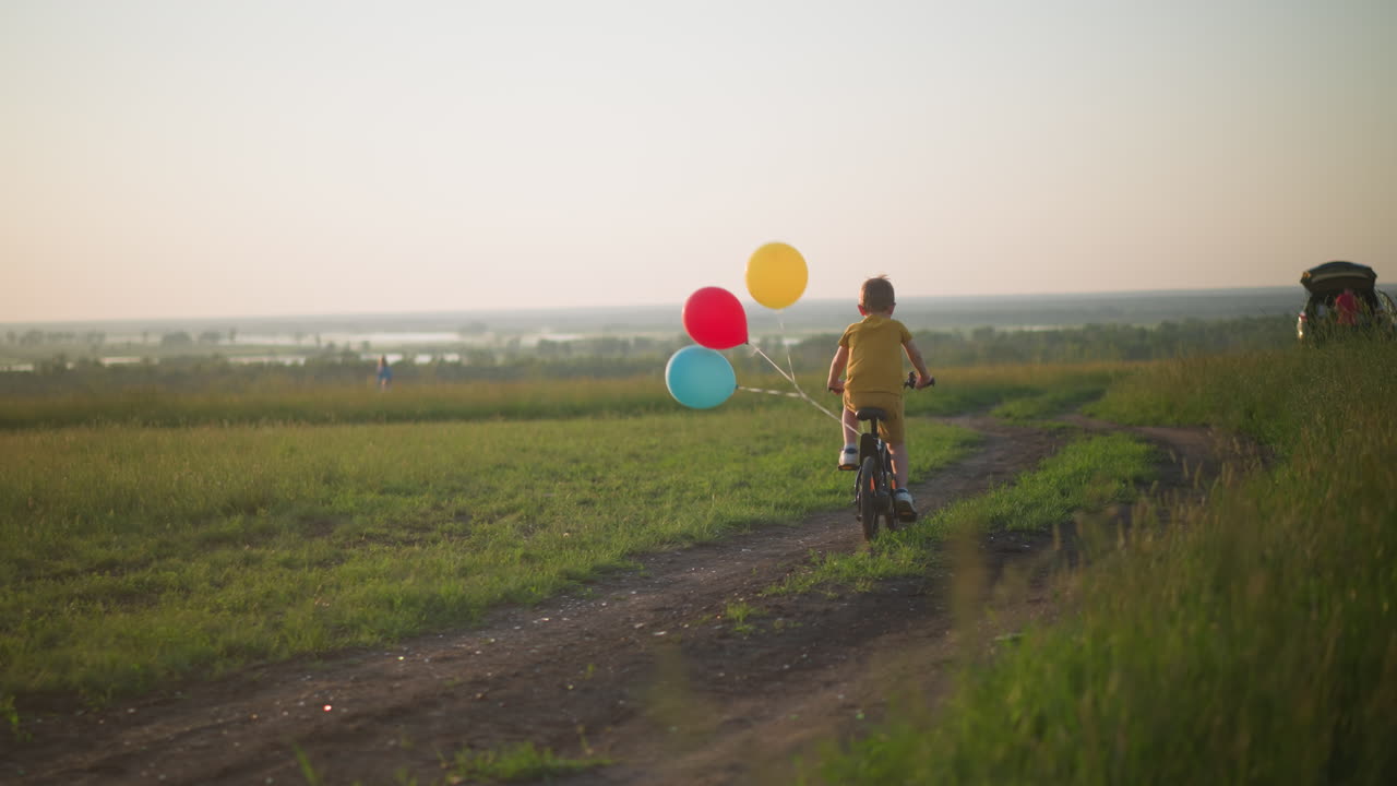 A young boy, dressed in yellow, rides his bicycle along a dirt path through a grassy field at sunset. Red, blue and yellow balloons are tied to the back of his bike. A car is visible in the background