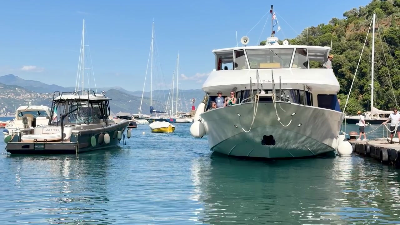 Un grande yacht bianco manovra lentamente verso un molo di pietra in un porto italiano dalle acque turchesi.