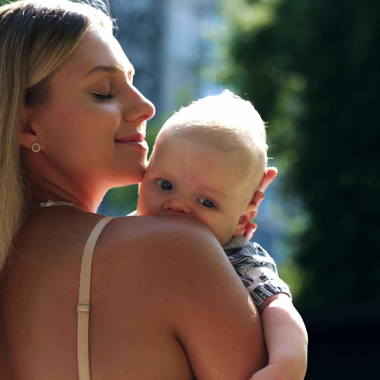 Beautiful blonde woman stands half-back to camera holding a baby. Loving mom caressing her son tenderly. Close up