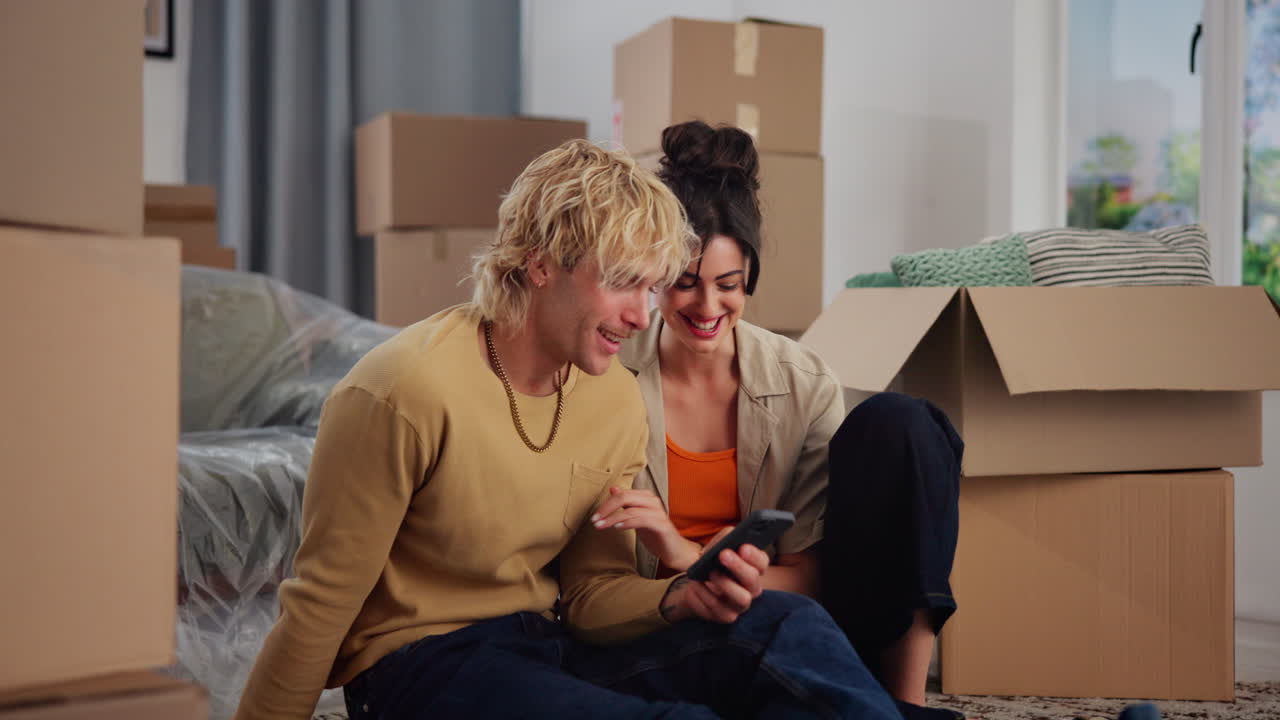 Happy Young Couple Sitting on Floor and Looking at Smartphone in New Home