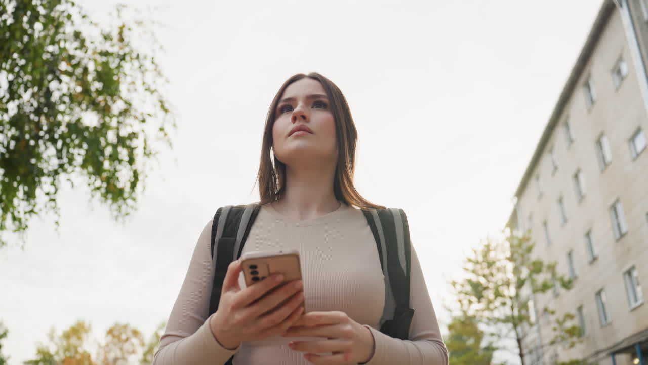 Giovane donna con uno zaino che controlla il telefono all'aperto, in una strada residenziale, inquadrature dal basso, condomini e alberi autunnali sullo sfondo, scorrendo l'app di consegna e in attesa dell'ordine, neutro