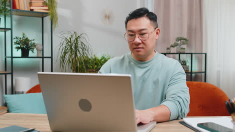 Asian-mature-man-freelancer-starts-working-on-laptop,-sends-messages,-makes-online-purchases-at-home