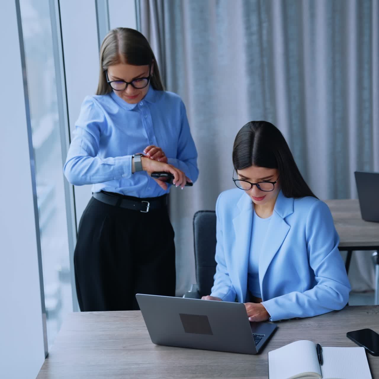 Female colleagues in office looking at laptop discussing work. Blonde woman standing and looking at her watch and then at computer again