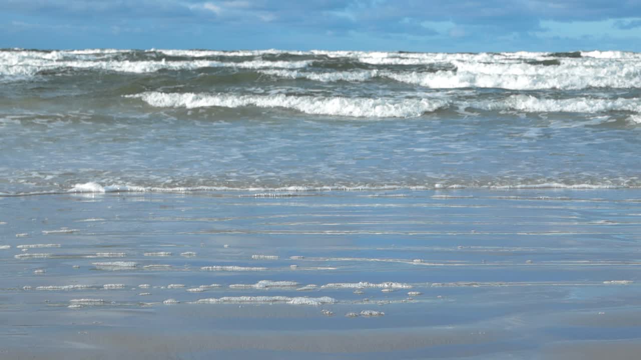Baltic ocean sea water waves coming to shore in slow motion with big waves, ripples and splashy white water leaving behind bubbles and foam on the sandy shoreline during day time in Vääna Jõesuu.