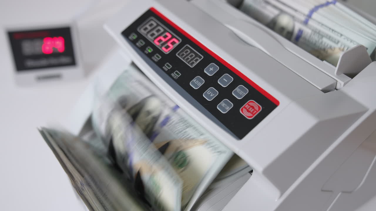 Male hand inserts a wad of hundred dollar bills into a slot of cash counting machine. Equipment for financial operations close up. Blurred backdrop.