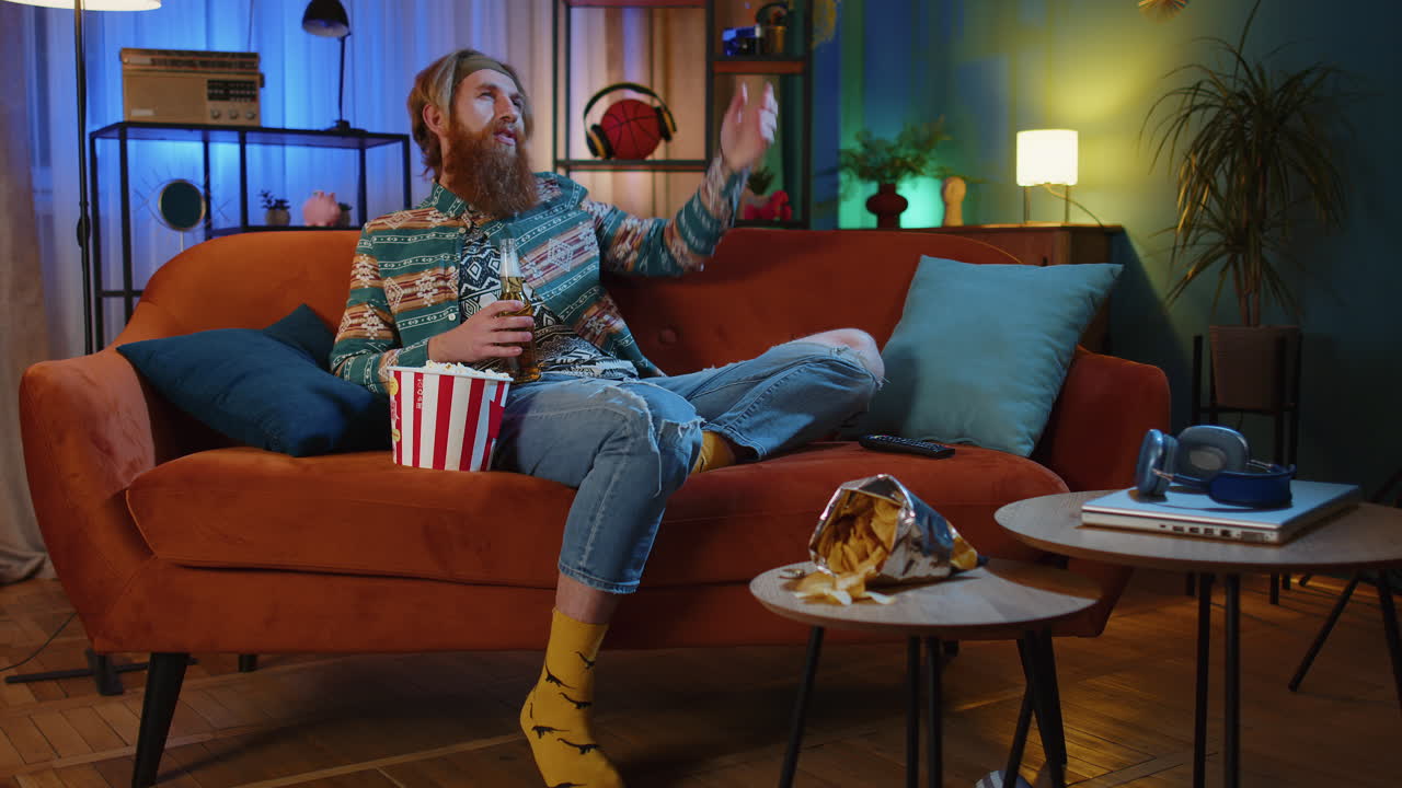 un hombre está sentado en el sofá de su sala de estar, comiendo palomitas de maíz y bebiendo cerveza mientras ve la televisión.