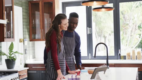 Feliz-Pareja-Diversa-Usando-Delantales-En-La-Cocina-Y-Usando-Tableta,-Cámara-Lenta