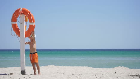 orange lifebuoy hanging from a pole on the beach. a curious boy on the beach stands next to a lifebuoy against the background of the azure sea