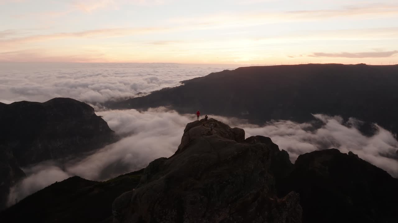 Drone footage of hikers standing on a rocky peak above dense cloud cover during sunrise at Pico do Arieiro in Madeira, Portugal.