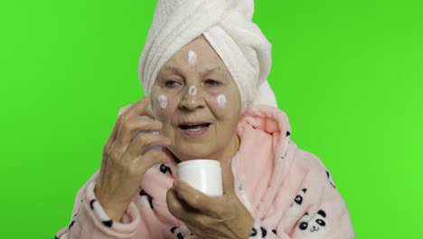 elderly grandmother in bathrobe. old woman applying moisturizing cream on face