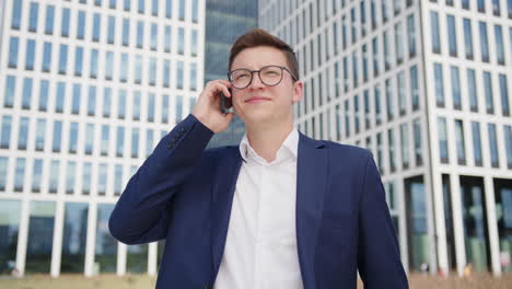 A-young-successful-entrepreneur-talks-on-the-phone-and-smiles-into-the-camera-in-front-of-the-modern-office-building