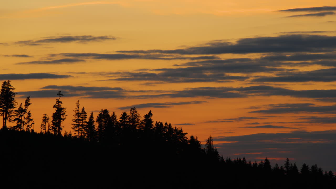 Zoome on the hilltops where dark trees are hidden in the background of shadows and silhouettes of clouds moving on the horizon during sunset
