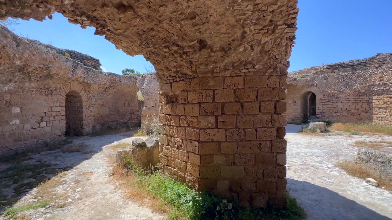 Archi e portali in pietra consumati dal tempo incorniciano un cortile aperto tra le rovine di Cartagine, con una forte luce di mezzogiorno e ombre profonde. Tunisia.