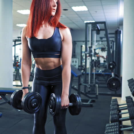 athletic young woman with perfect body in sportswear lifting dumbbells in the gym. red-haired female is doing her muscles with dumbbells during training on the background of sports club.