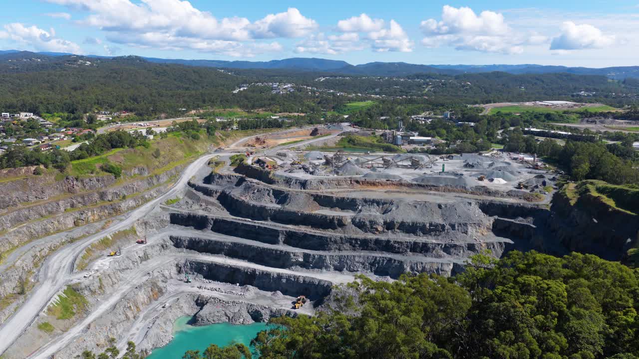 Drone footage captures a vast quarry with terraced levels under bright daylight, surrounded by lush greenery and distant hills