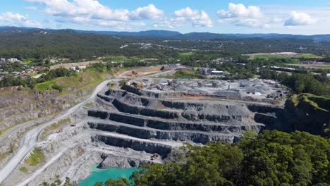 drone footage captures a vast quarry with terraced levels under bright daylight, surrounded by lush greenery and distant hills