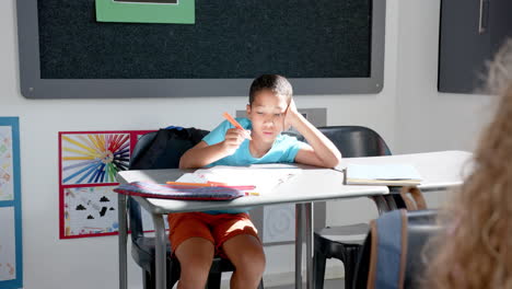biracial boy focused on writing in a classroom, with a pencil in hand and books on the desk