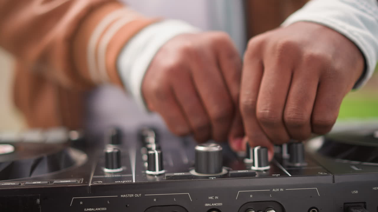 Gros plan sur les mains d'un DJ noir mixant sur un contrôleur, scène de rue avec un bokeh chaleureux, réglages tactiles des potentiomètres et faders, manches de veste décontractée, set en extérieur en journée, ambiance entraînante immersive et concentration.
