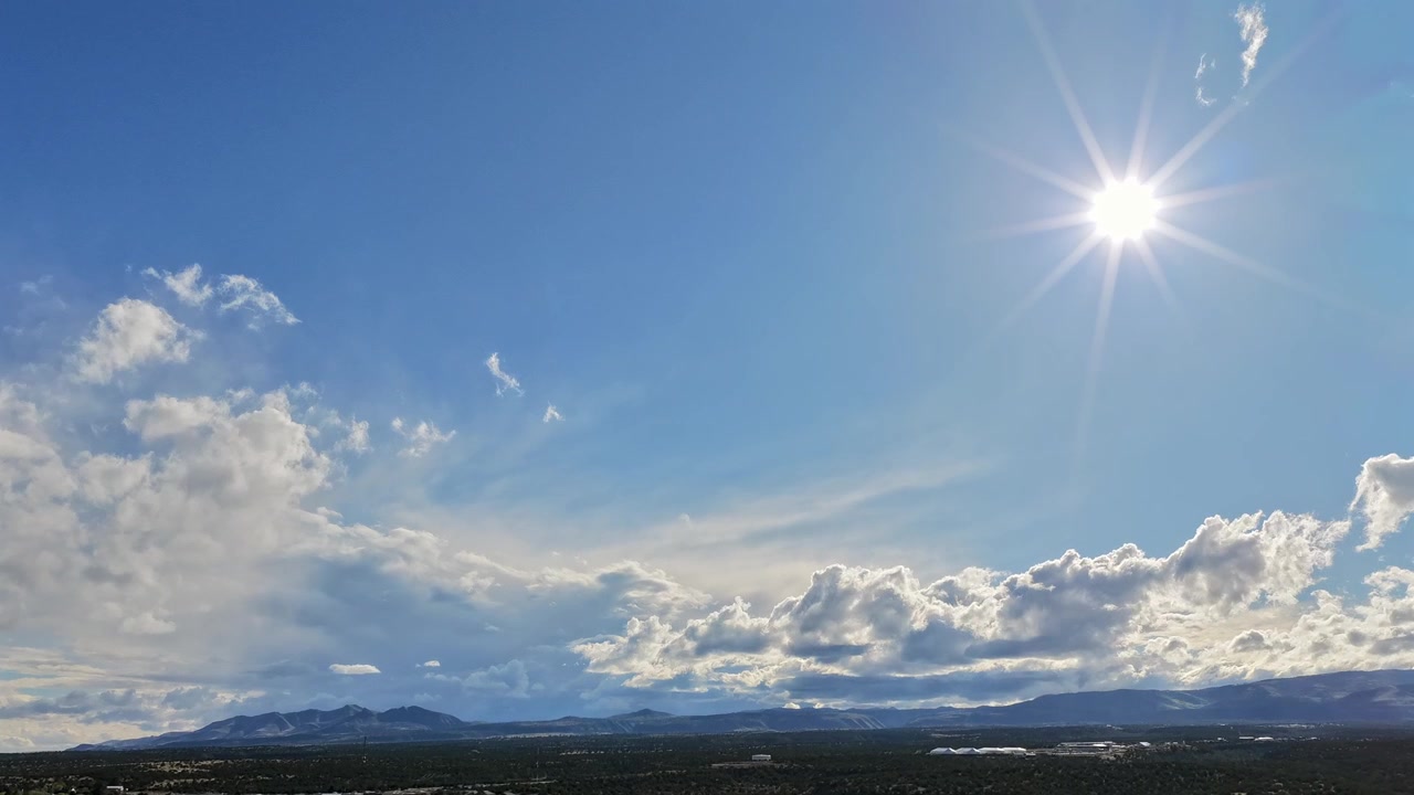 Un timelapse 4K de dron captura un cielo azul brillante con movimientos de nubes blancas sobre escarpados picos montañosos. El movimiento cinematográfico revela capas cambiantes y luz solar brillante sobre un vasto paisaje de gran altitud.