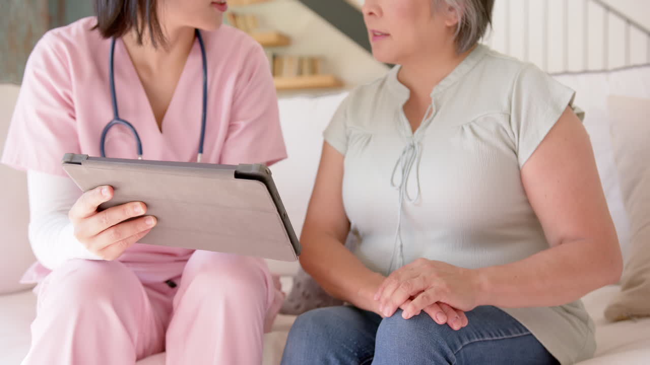 Nurse in pink scrubs showing senior asian woman information on tablet during home visit