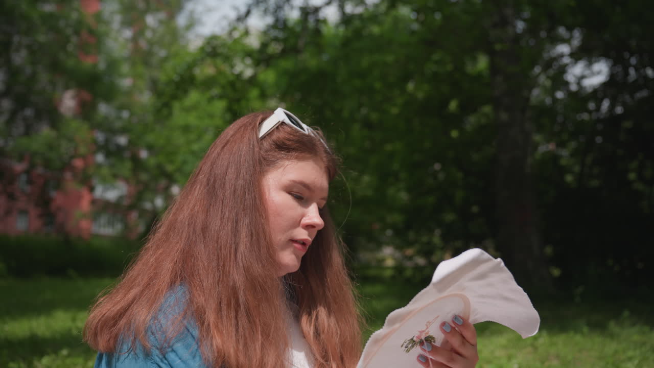 Close up of student concentrating on embroidery hoop while seated outdoors near residential building, creating colorful pattern under sunlight, showing patience